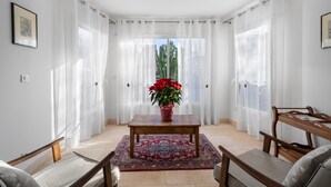 Ein Zimmer mit einem Holztisch, einem roten Teppich mit Muster, einer Topfpflanze mit roten Blüten und Blick durch transparente Vorhänge.