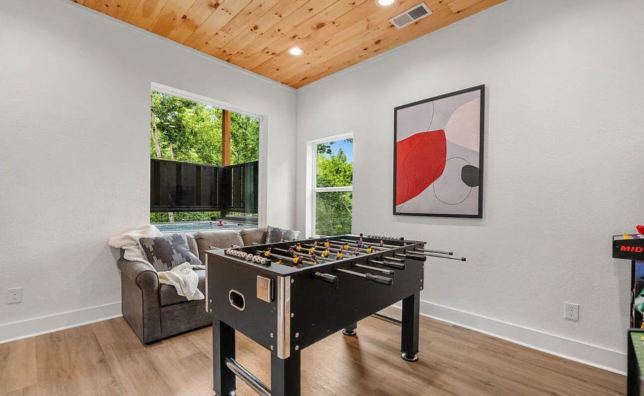 A room with a foosball table, a sofa, and a window with a view of greenery.