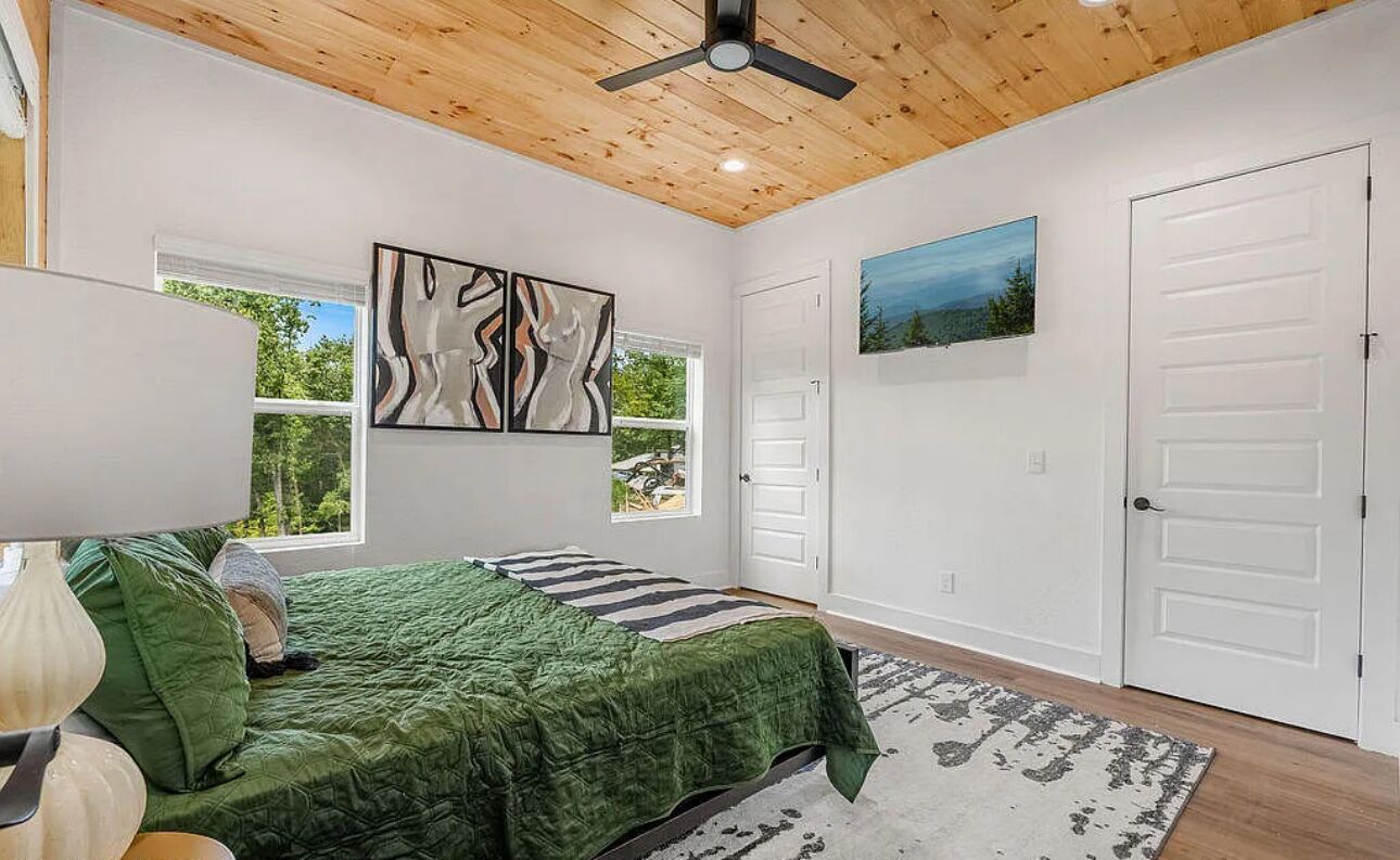 A bedroom with a bed, a ceiling fan, a painting, a window, and a door.
