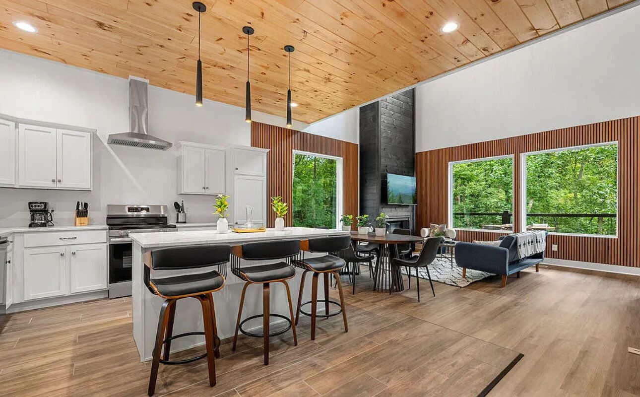 A modern kitchen with white cabinets, a central island with bar stools, a dining area with a sofa, and large windows with a view of greenery.
