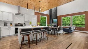 Eine moderne Küche mit weißen Schränken, einer zentralen Kochinsel mit Barhocker, einem Essbereich mit einer Couch und großen Fenstern mit Blick auf Grünflächen.