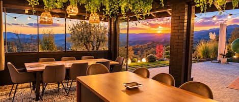 A dining area with a view of a sunset over mountains, featuring hanging plants, wicker lamps, and a brick wall.