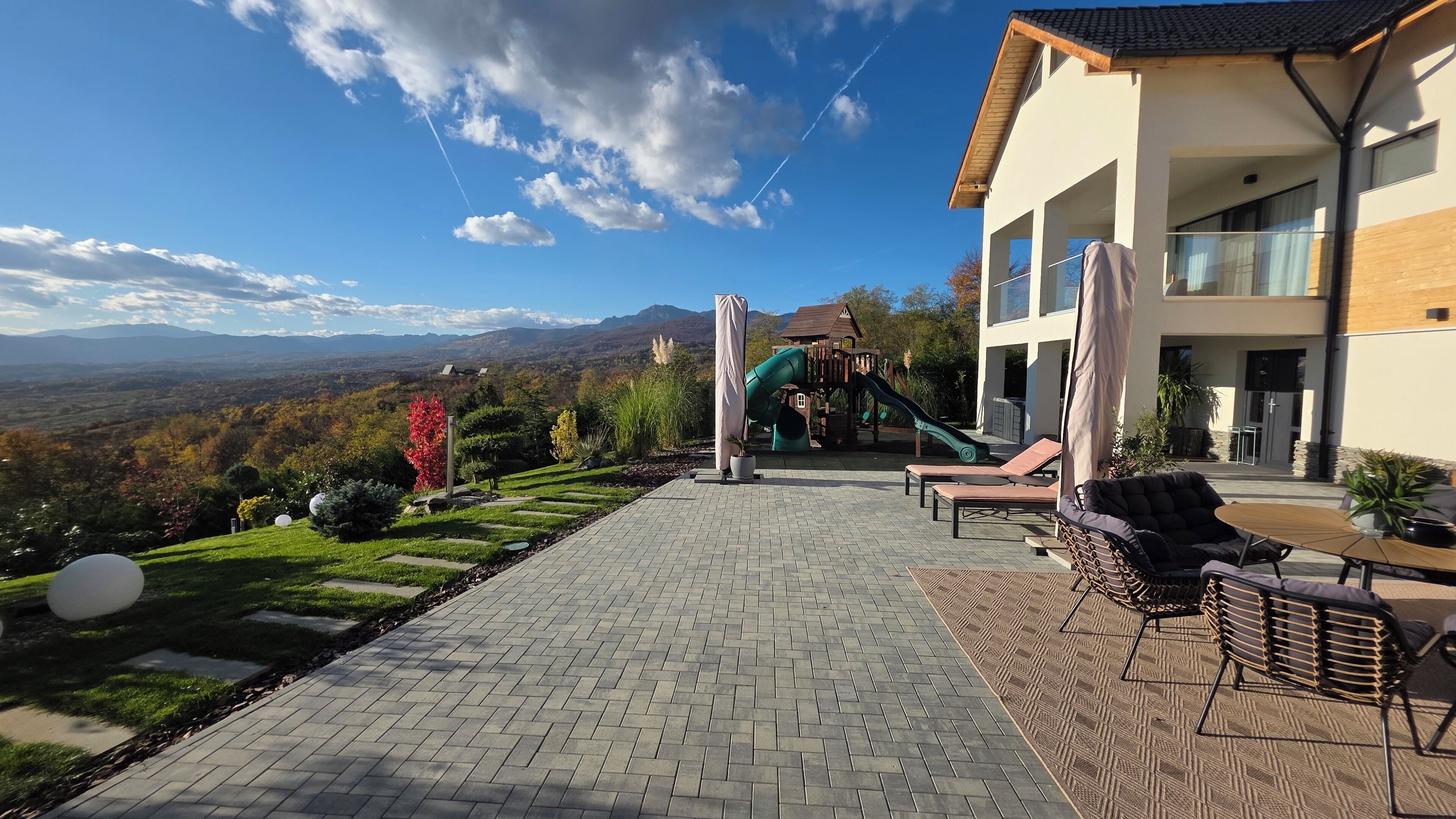 Un patio con suelo de ladrillo, mobiliario exterior, un tobogán y vistas a un paisaje montañoso.