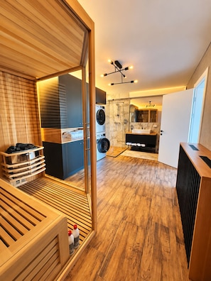 A modern bathroom with a glass shower enclosure, a built-in washing machine, and a wooden sauna bench.