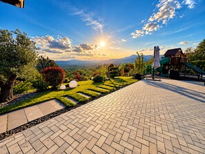 A paved outdoor area with a playground, including a slide and climbing structures, surrounded by landscaped gardens and a view of distant mountains.