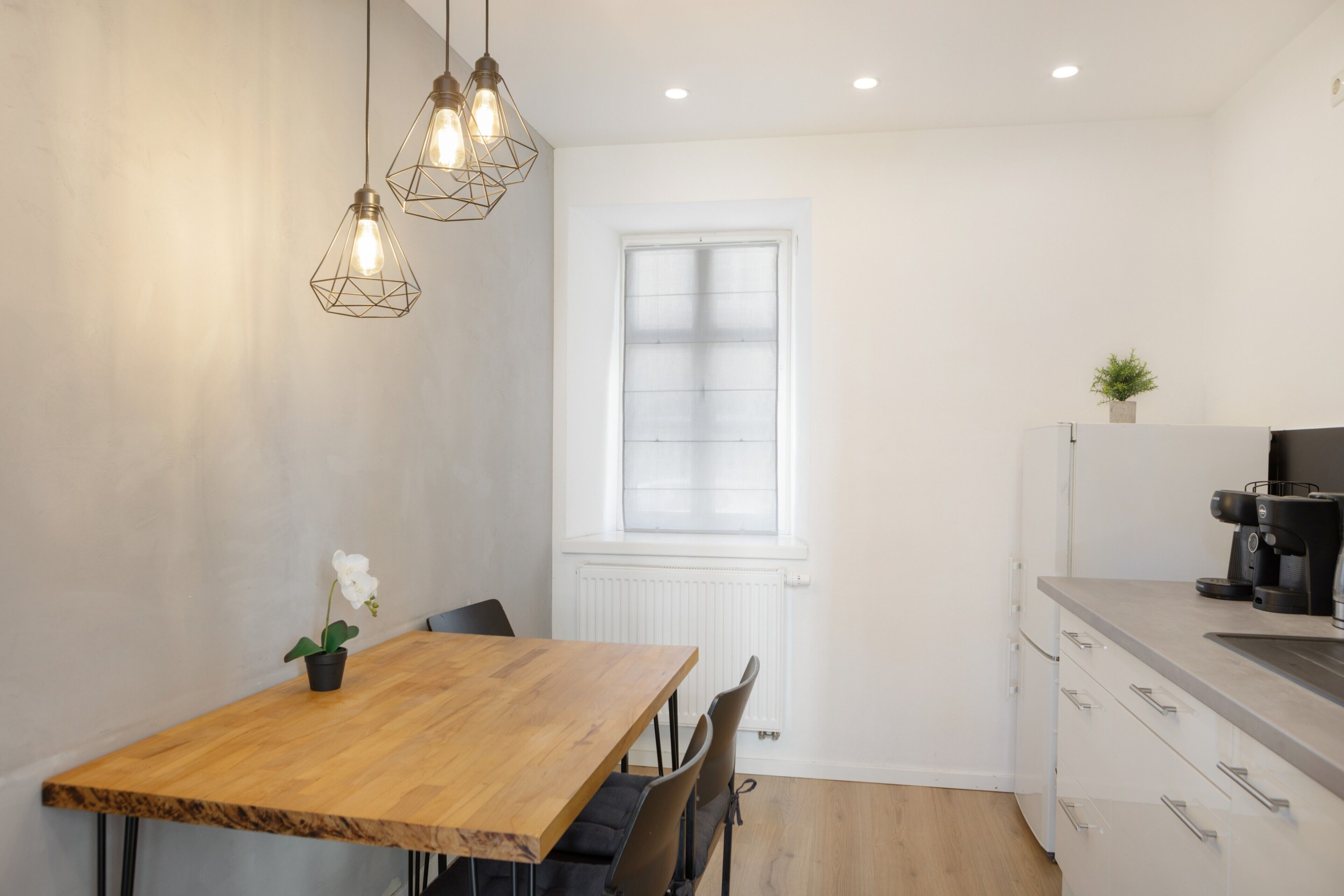 Eine moderne Küche mit Holztisch, Stühlen, Pendelleuchten, einem Fenster und einer Kaffeemaschine.