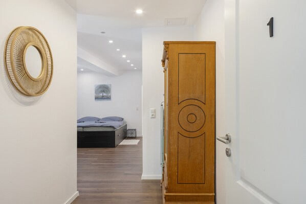 A hallway leading to a bedroom with a bed, a wooden door with a number, and a wall-mounted circular mirror.