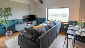 A modern living room with a sectional sofa, a flat-screen TV, a dining table set for two, and a large window offering a view of the countryside. - Black Bird Cottage (Gaerwen)