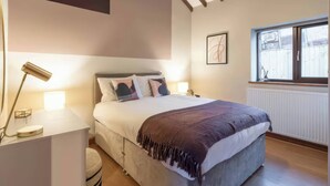 A bedroom with a bed, bedside lamps, a window, a radiator, and a ceiling light. - Black Bird Cottage (Gaerwen)