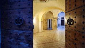 Um corredor interno com piso de azulejos, portas de madeira com detalhes metálicos e colunas de pedra.