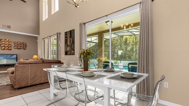 A dining area with a white table set for four, clear chairs, and a large window with curtains. - Upstay Home w Pool Hot Tub Patio - Pet Friendly (Kissimmee)