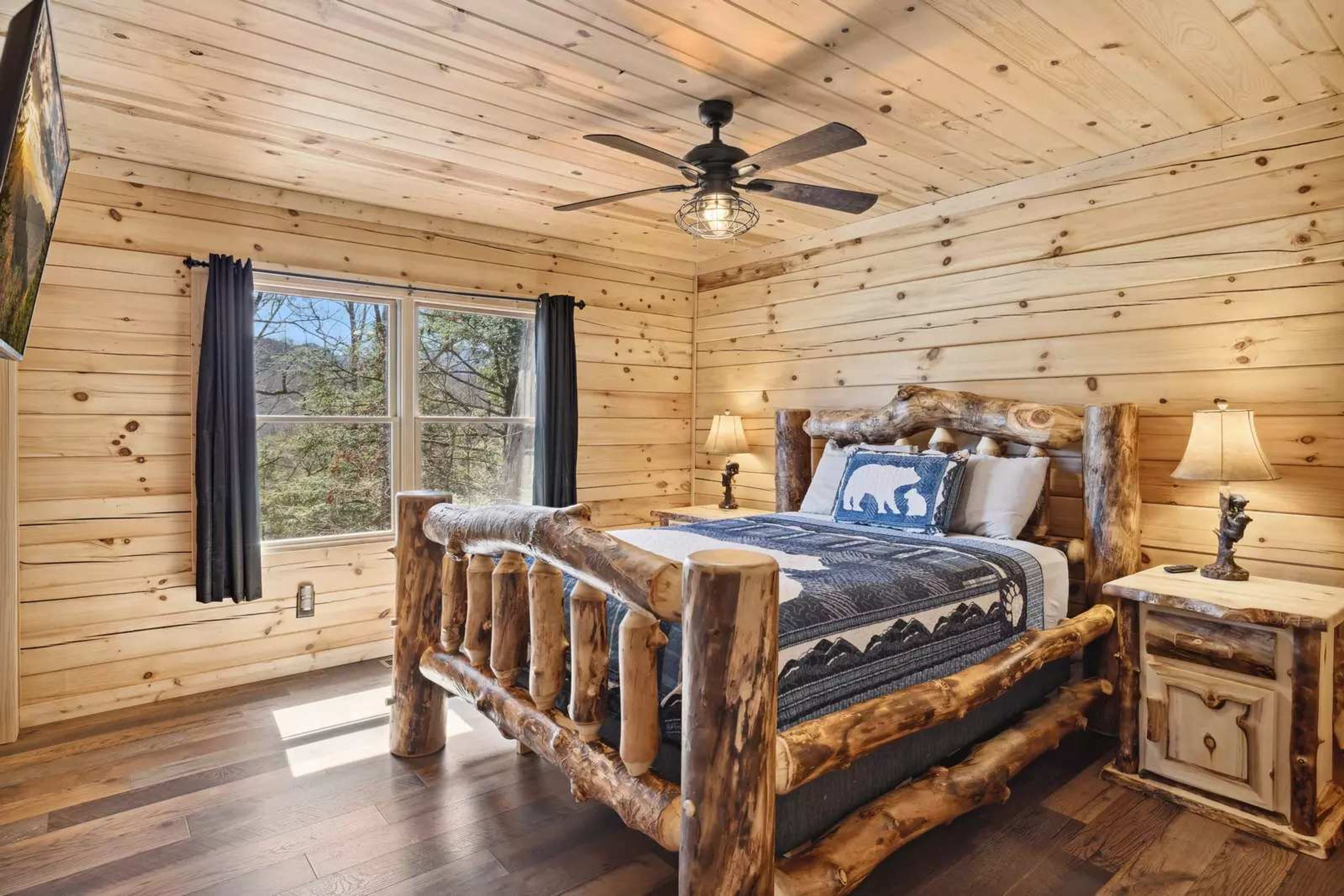 Un dormitorio de cabaña de madera con una cama de madera, un ventilador de techo, una mesita de noche y una ventana con cortinas.