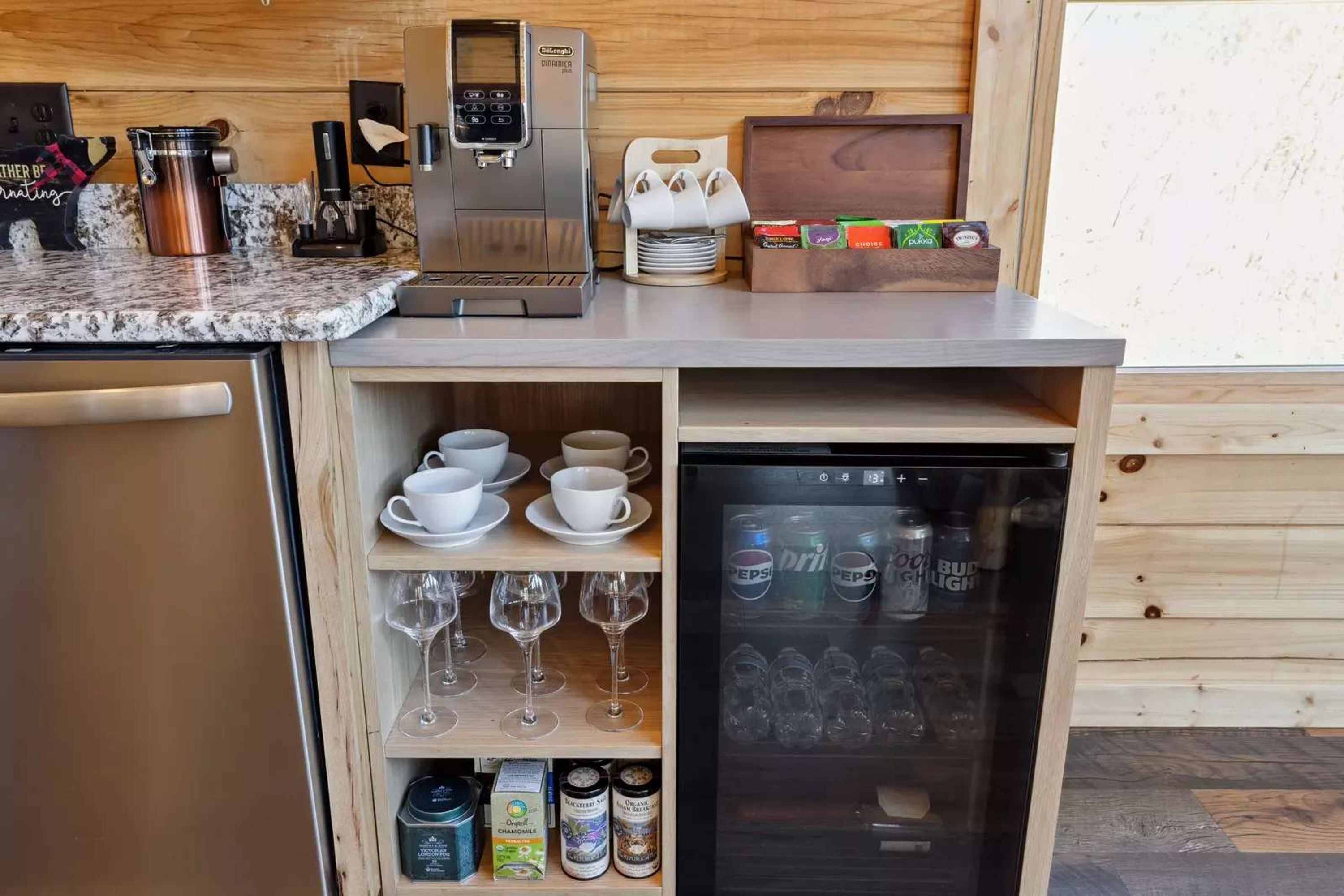 A kitchenette with a coffee machine, a mini-fridge stocked with beverages, and shelves with cups and wine glasses.