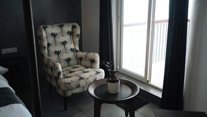 Um quarto com uma poltrona de estampa floral, uma mesinha com um vaso de planta e vista para uma varanda através de uma grande janela.