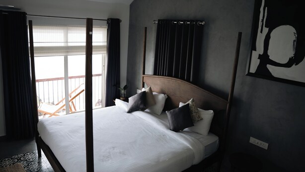 Quarto com cama grande, cortinas escuras, cabeceira de madeira e vista para varanda.
