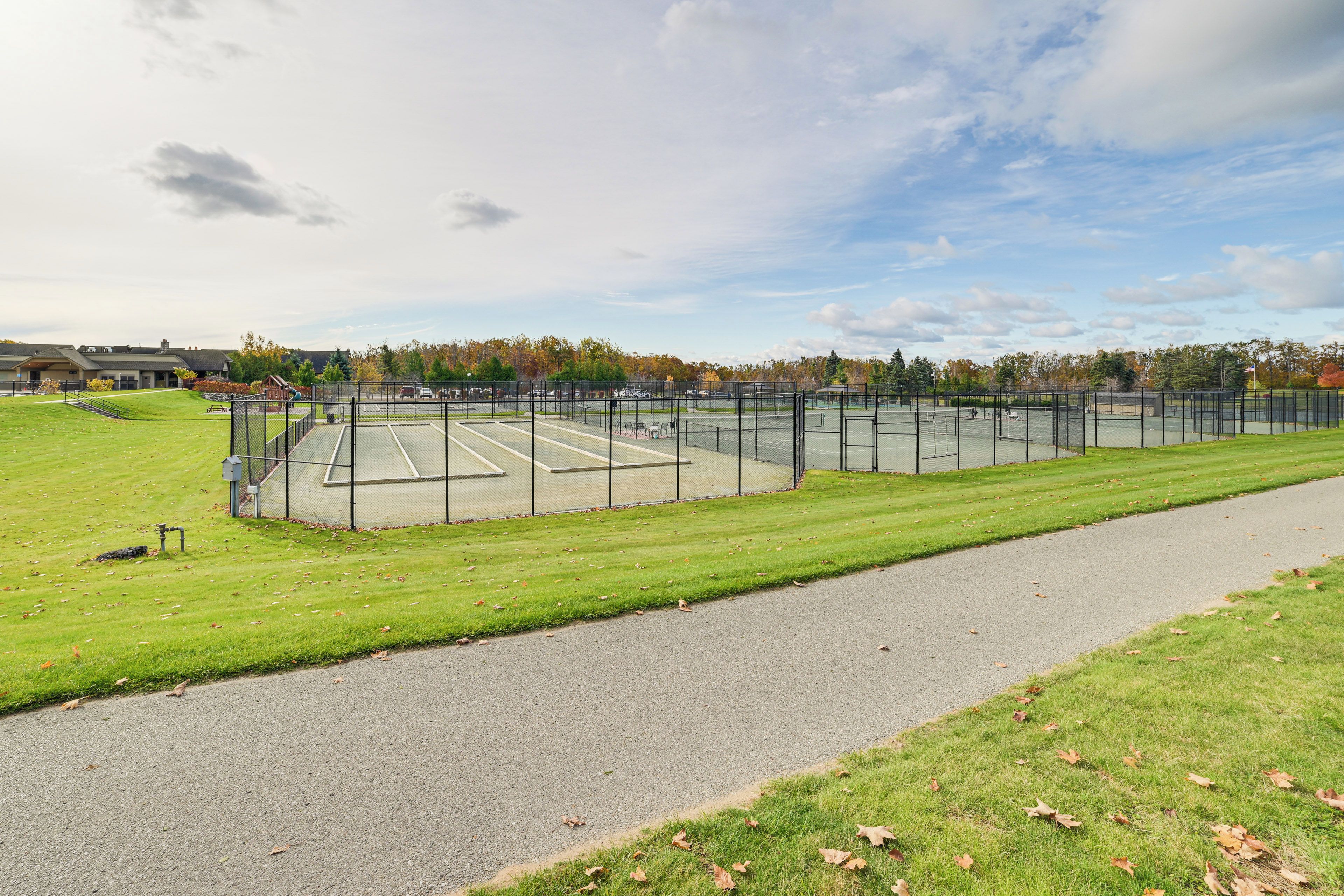 A tennis court with a surrounding fence, a paved walkway, and a grassy area.