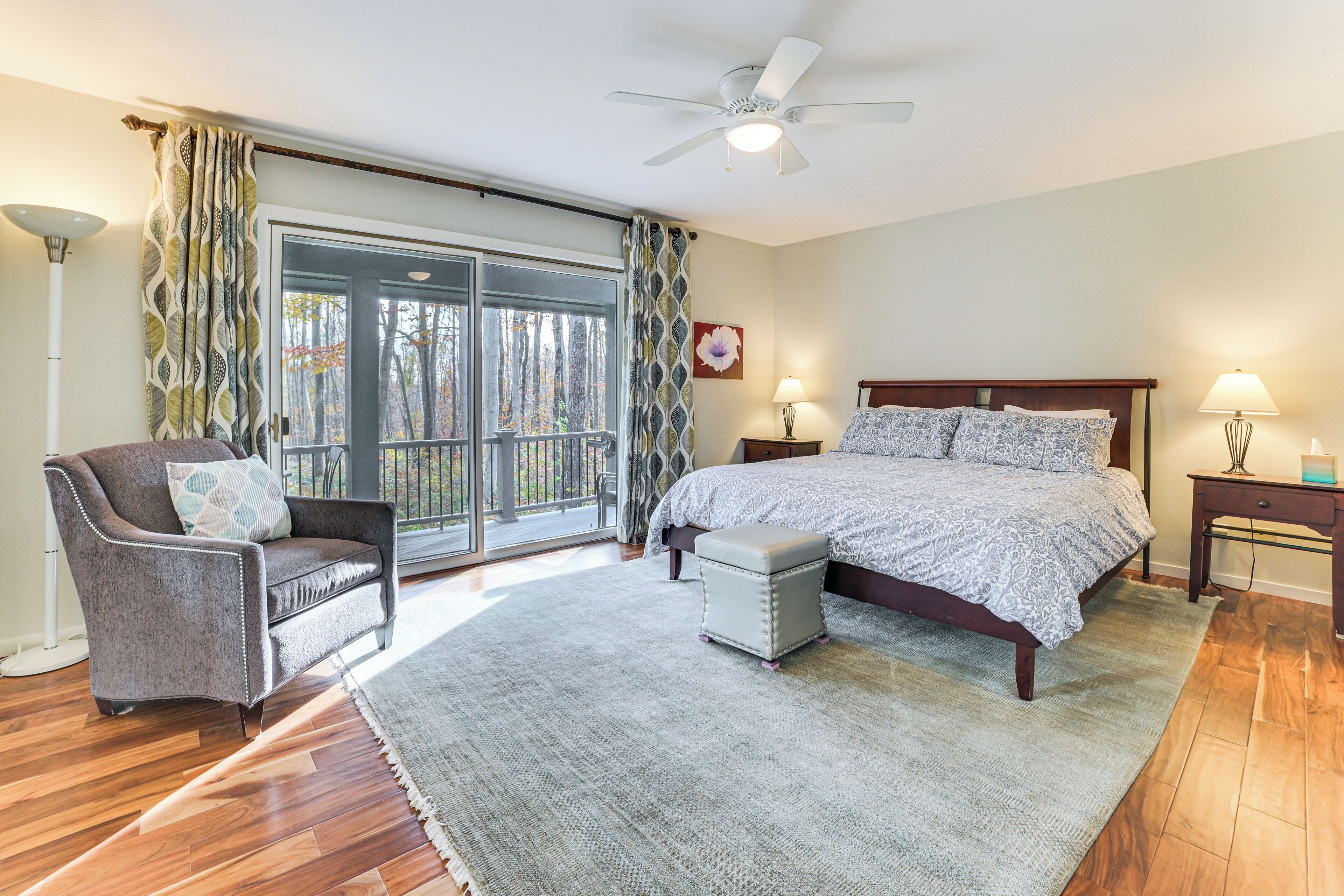 A bedroom with a bed, a chair, a nightstand, a lamp, a ceiling fan, and a view through a glass door.