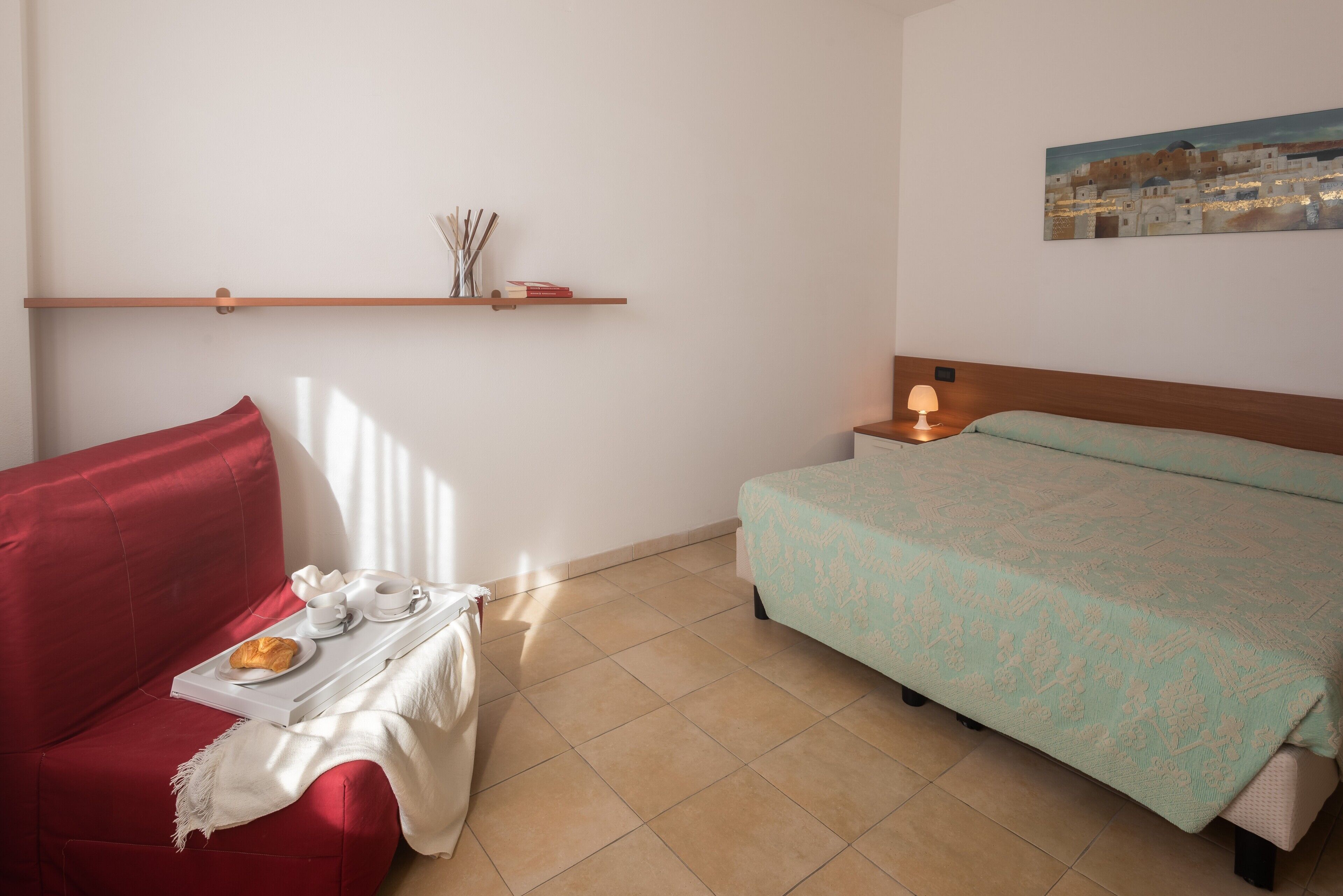 A room with a red sofa, a bed with a patterned bedspread, a bedside table with a lamp, and a wall-mounted shelf with books and decorative items.