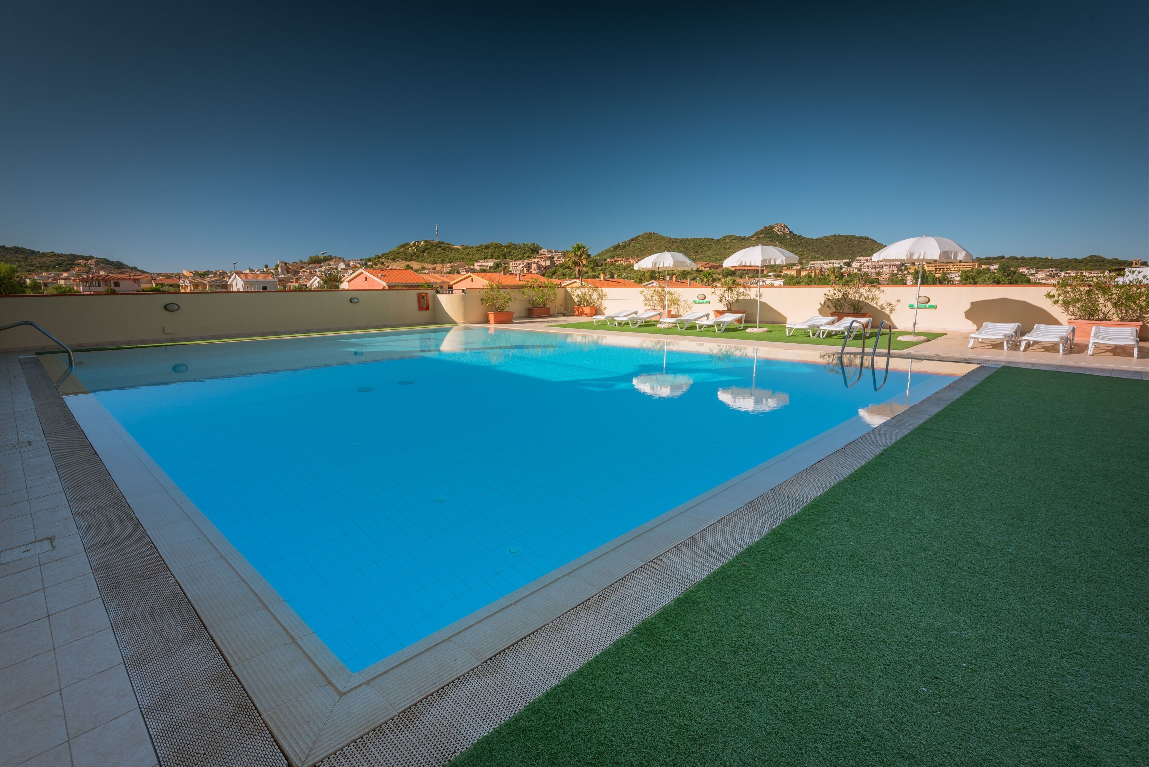A swimming pool with a clear blue water, surrounded by a green lawn and white lounge chairs.