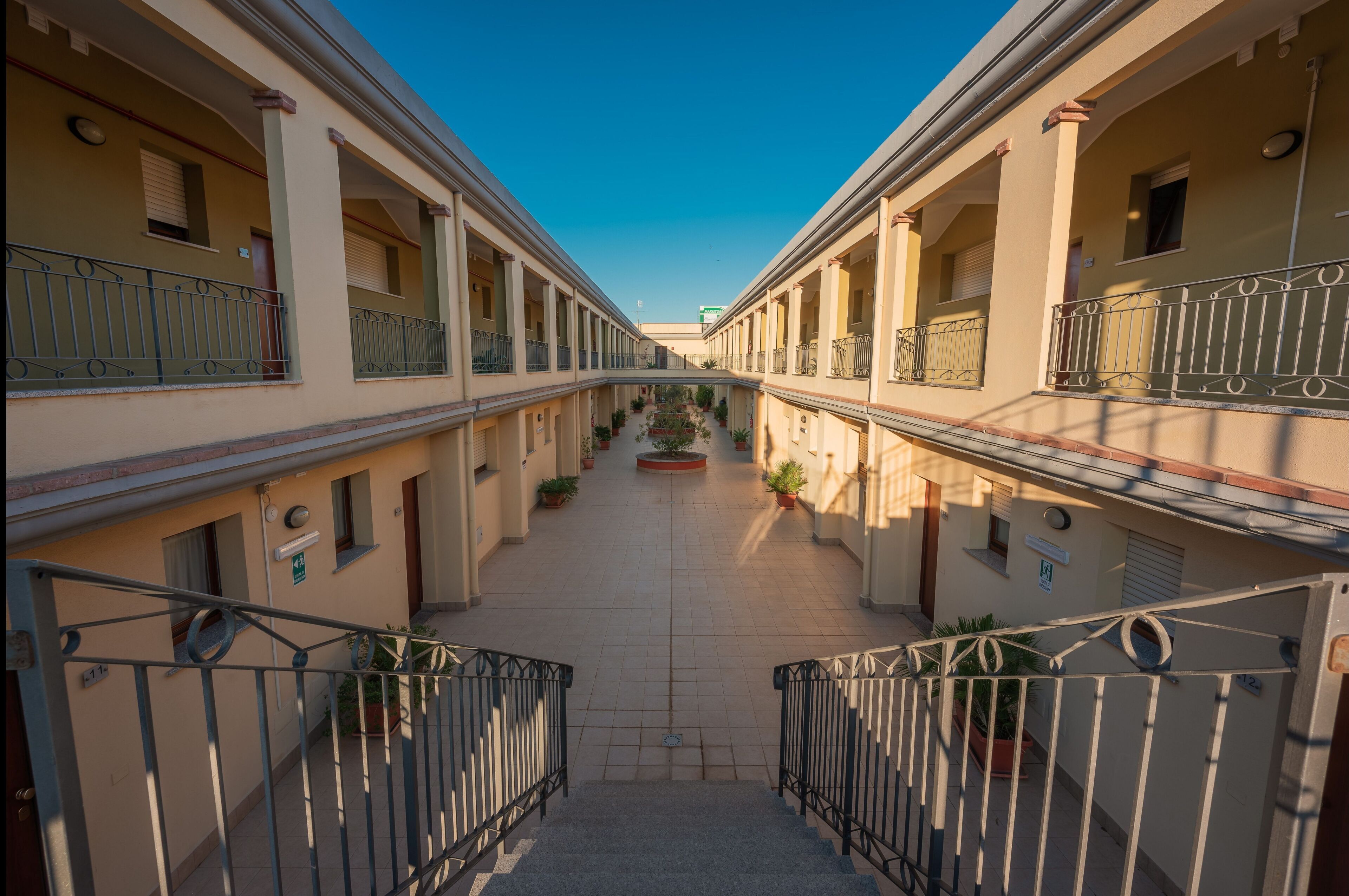 A courtyard with a central pathway flanked by buildings with balconies and potted plants.