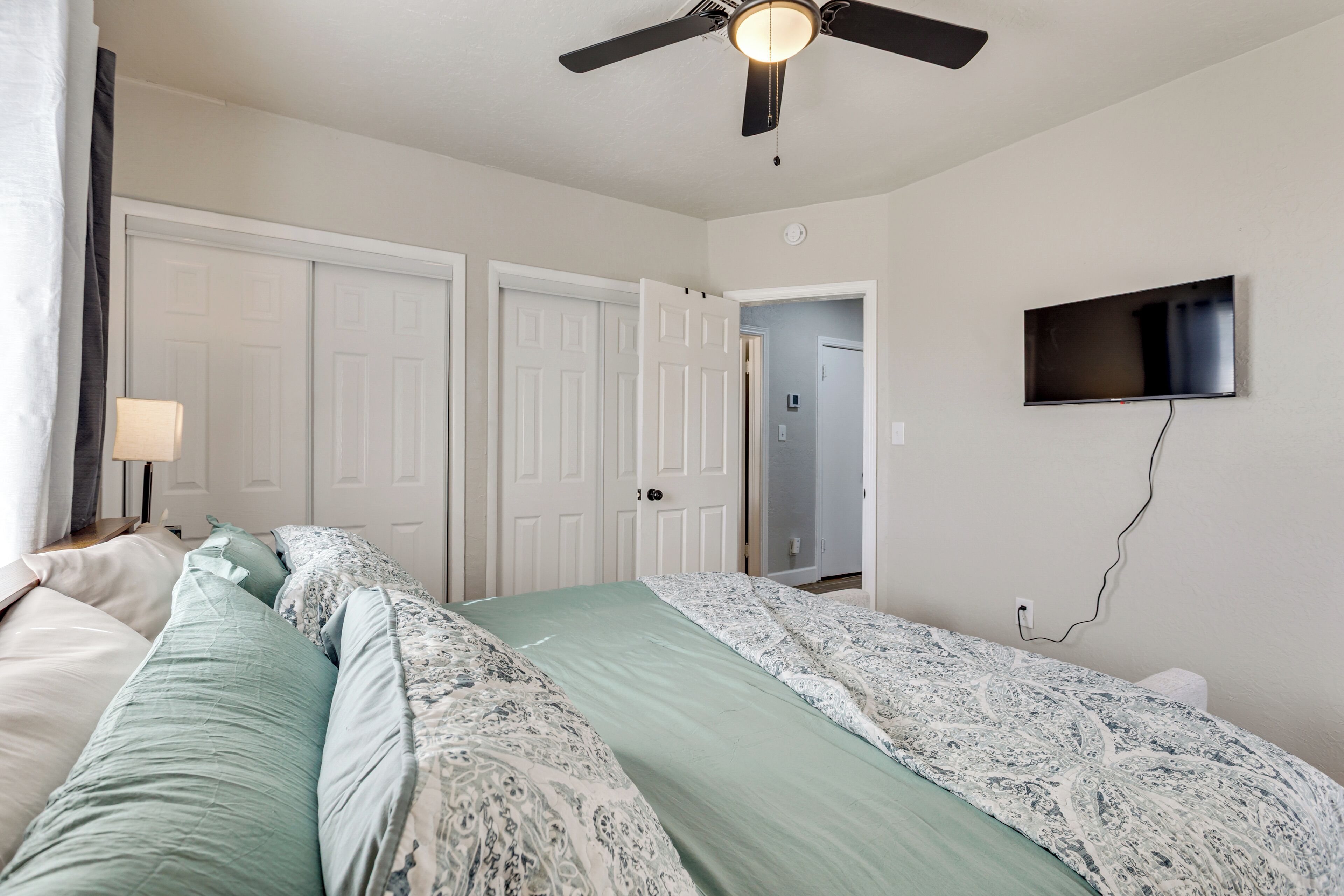 A bedroom with a bed, a ceiling fan, a TV mounted on the wall, and a door leading to another room.