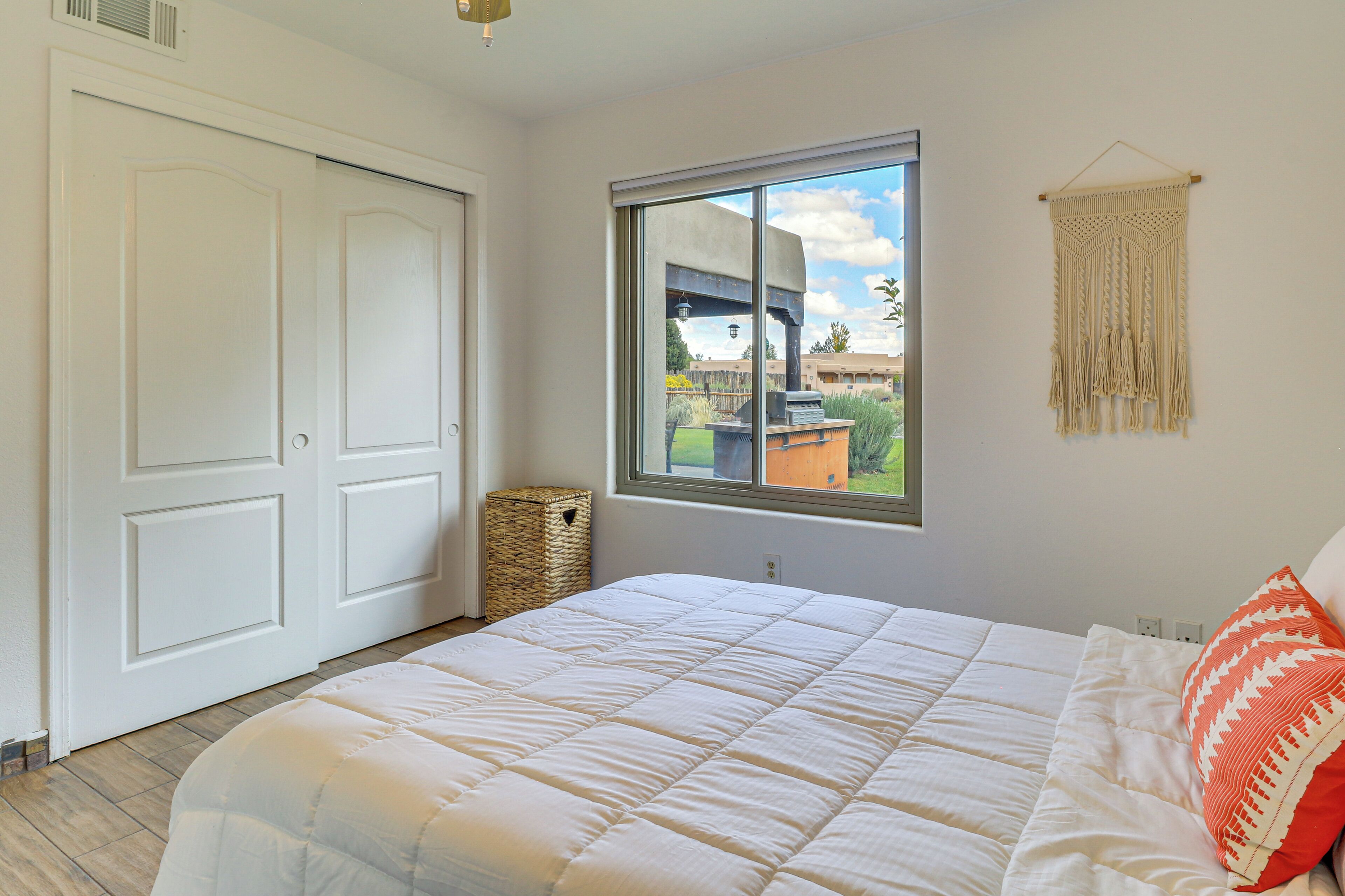 A bedroom with a large bed, a window with a view of the outdoors, a woven basket, and a macrame wall hanging.
