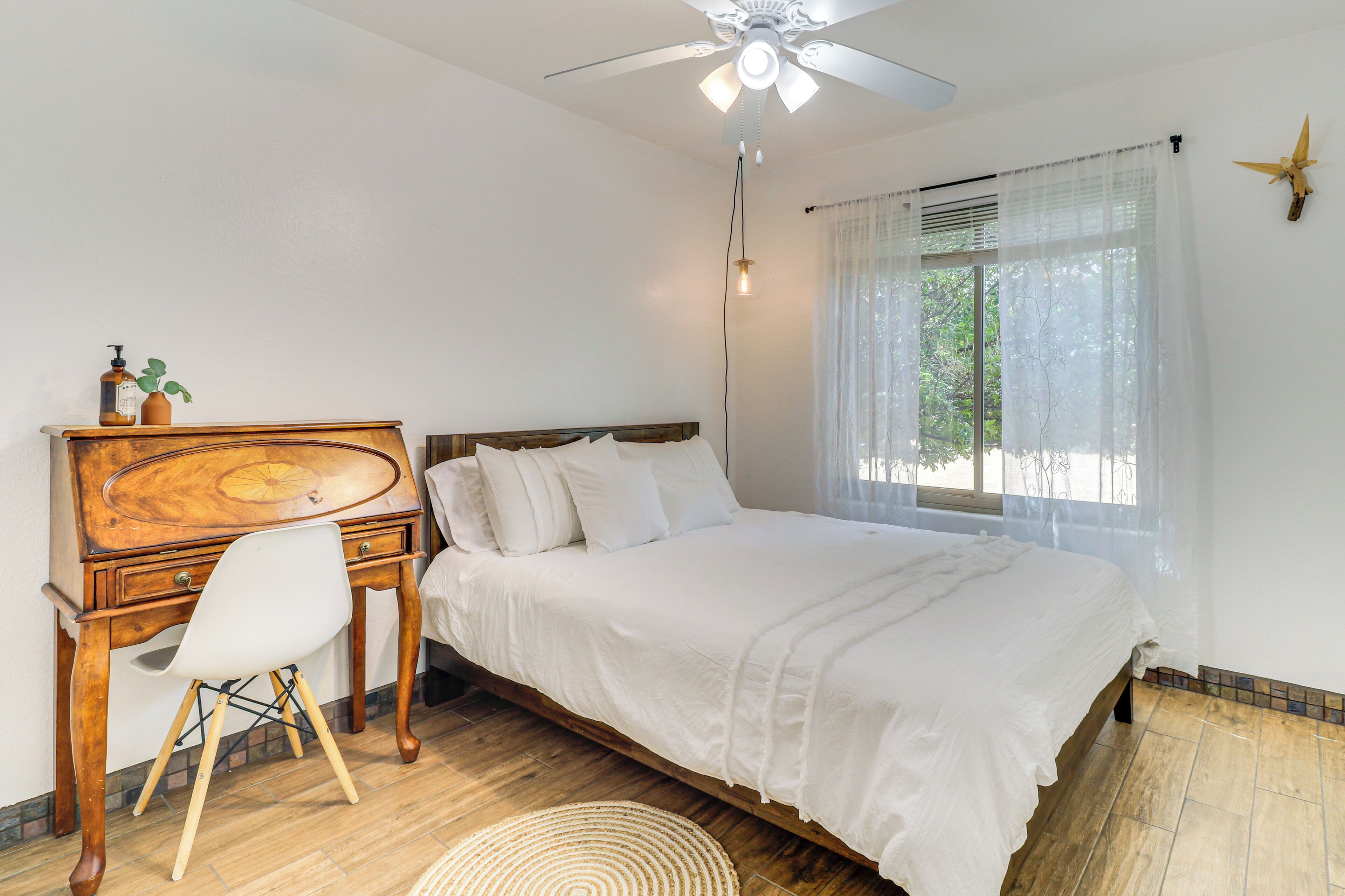 A bedroom with a wooden desk, a white chair, a bed with white bedding, a ceiling fan, and a window with sheer curtains.
