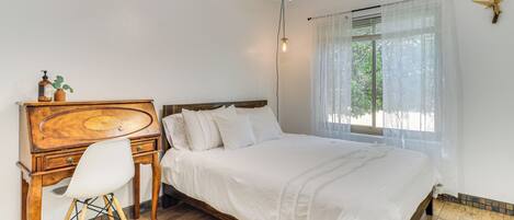 A bedroom with a wooden desk, a white chair, a bed with white bedding, a ceiling fan, and a window with sheer curtains.