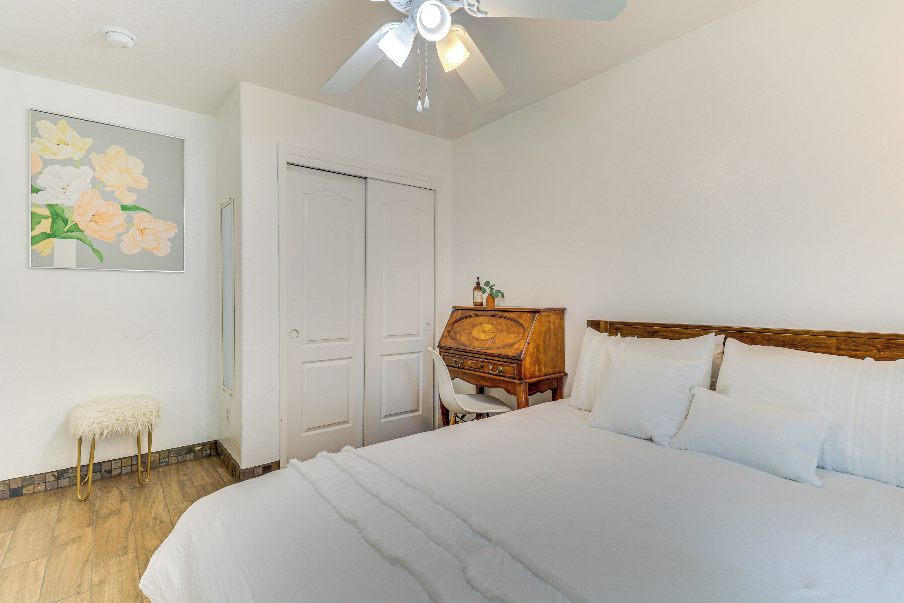 A neatly arranged bedroom with a bed, a wooden nightstand, a chair, a small stool, a painting, and a ceiling fan.