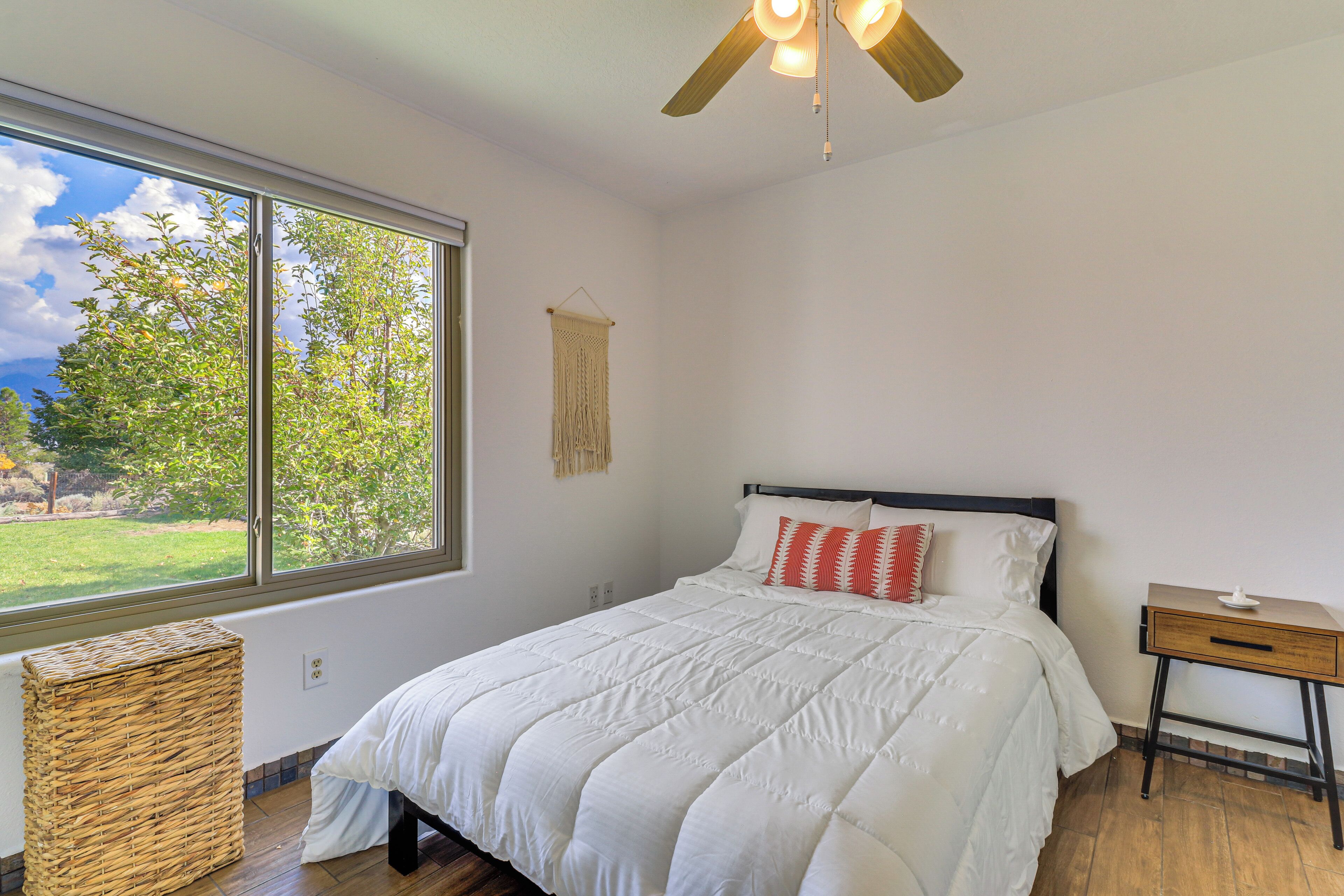 A bedroom with a large window, a bed with white bedding, a wooden nightstand, and a ceiling fan.