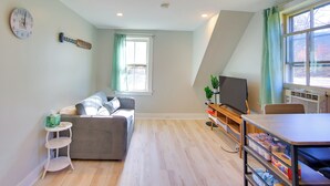 Una sala de estar moderna con un sofá gris, una mesita blanca, un mueble bajo para televisión de madera y un amplio ventanal con cortinas.