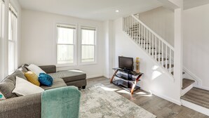 A spacious living room with a grey sofa, ottoman, and a television on a wooden stand. A staircase with white railings leads to the upper floor. - Private Yard! East Jersey Hub By River & Soccer! (National Park)