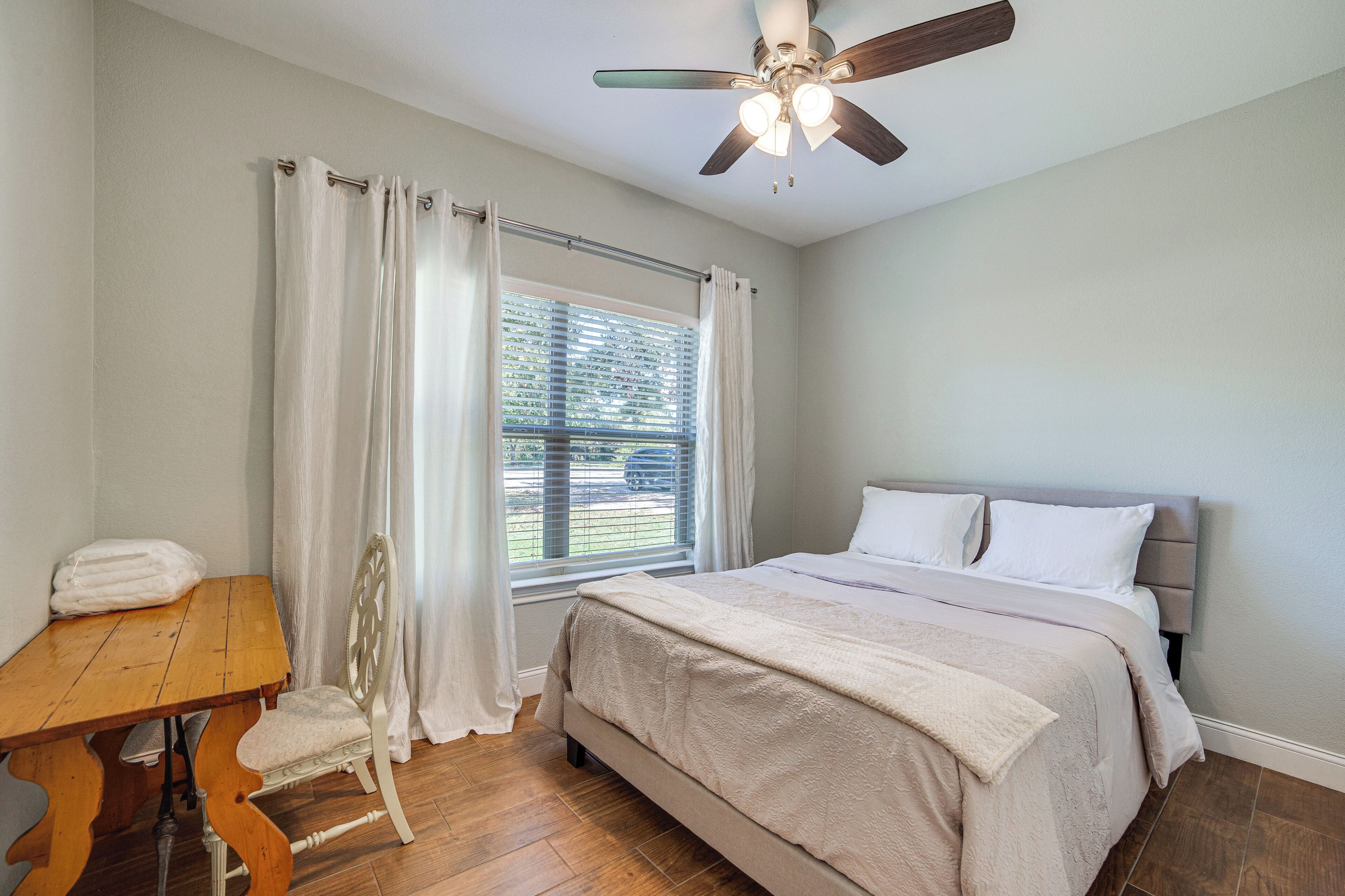 A bedroom with a bed, a wooden table, a chair, a window with curtains, and a ceiling fan.