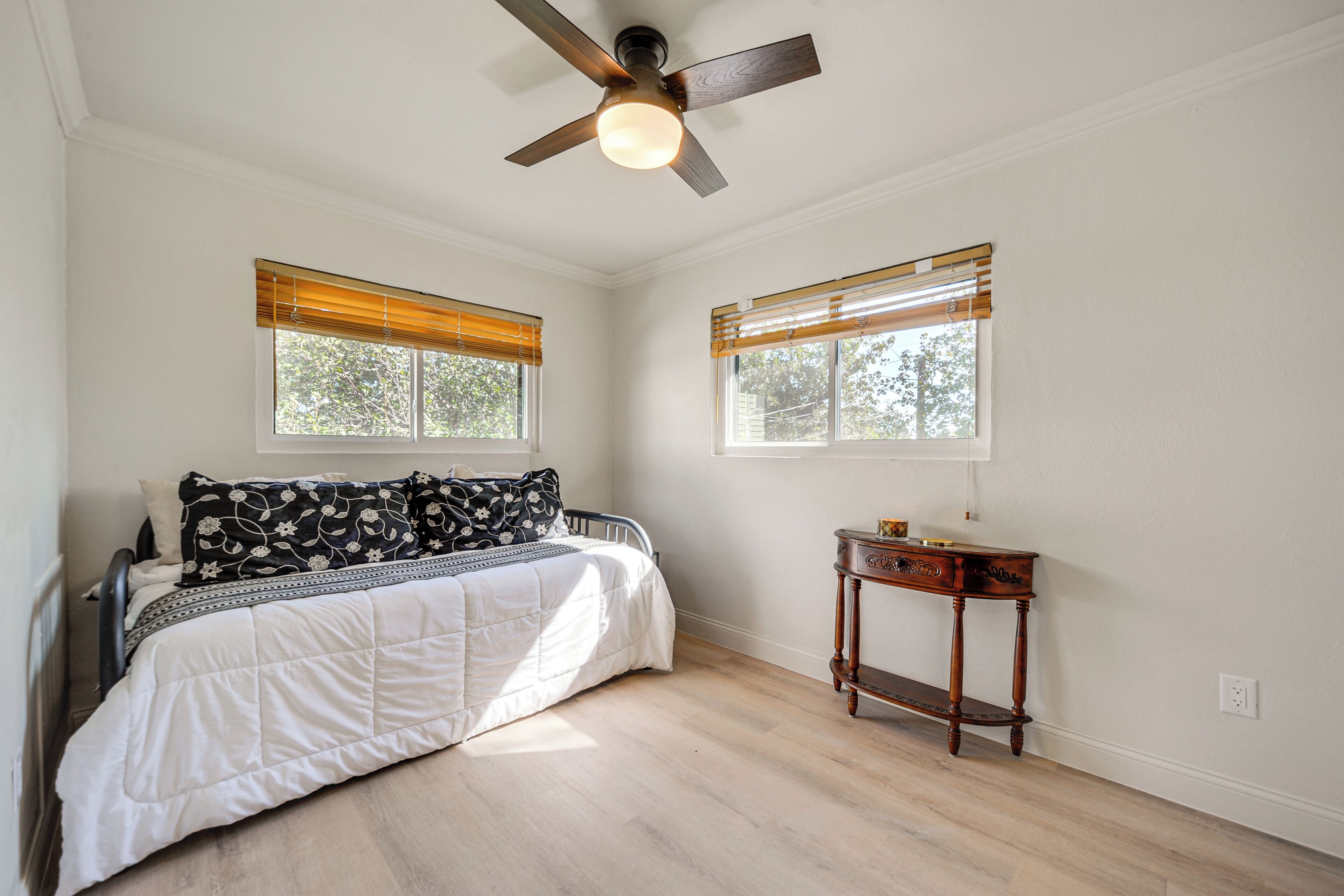 A bedroom with a bed, a wooden nightstand, a ceiling fan, and two windows with blinds.