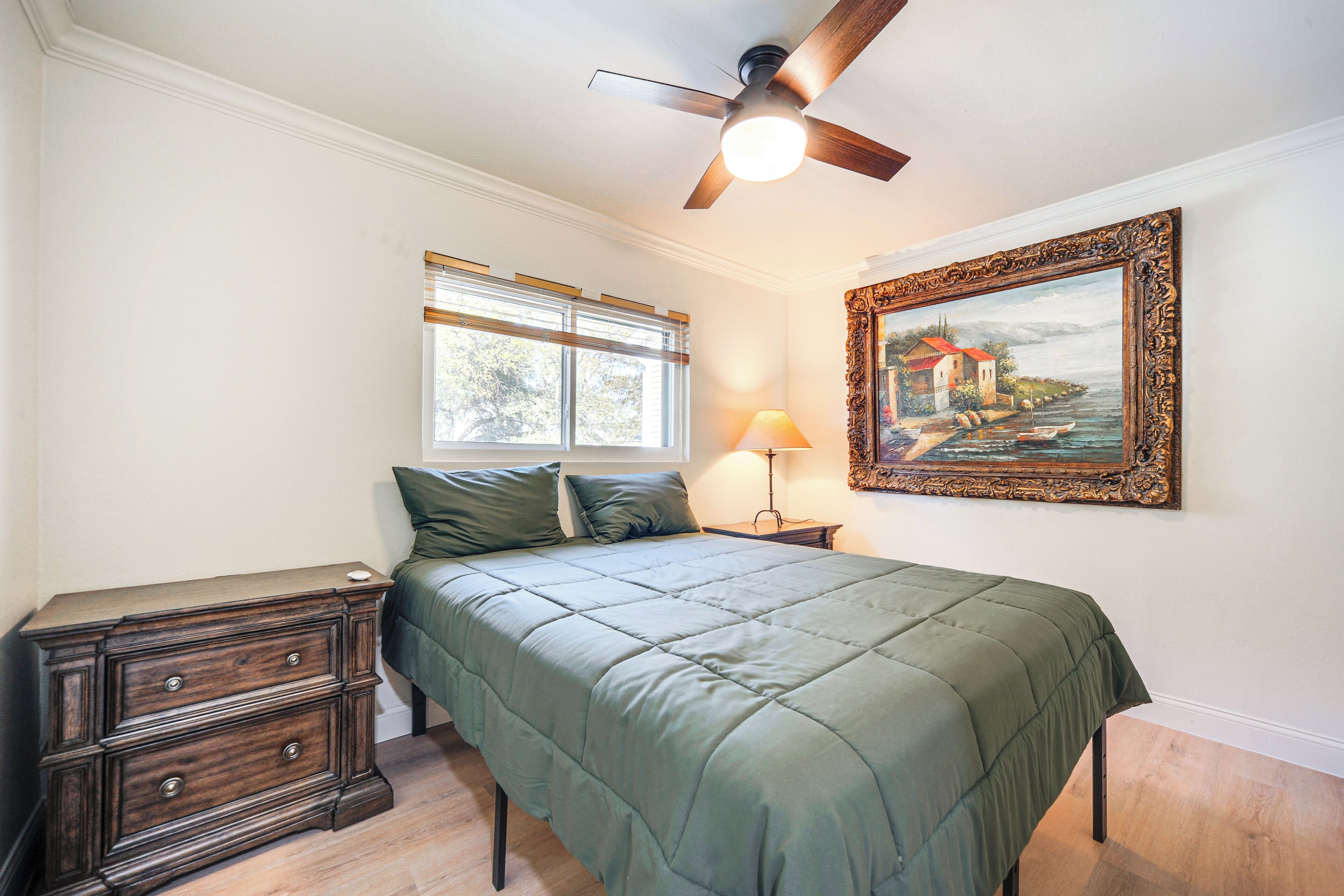 A bedroom with a bed, a dresser, a ceiling fan, a window with blinds, and a framed painting on the wall.