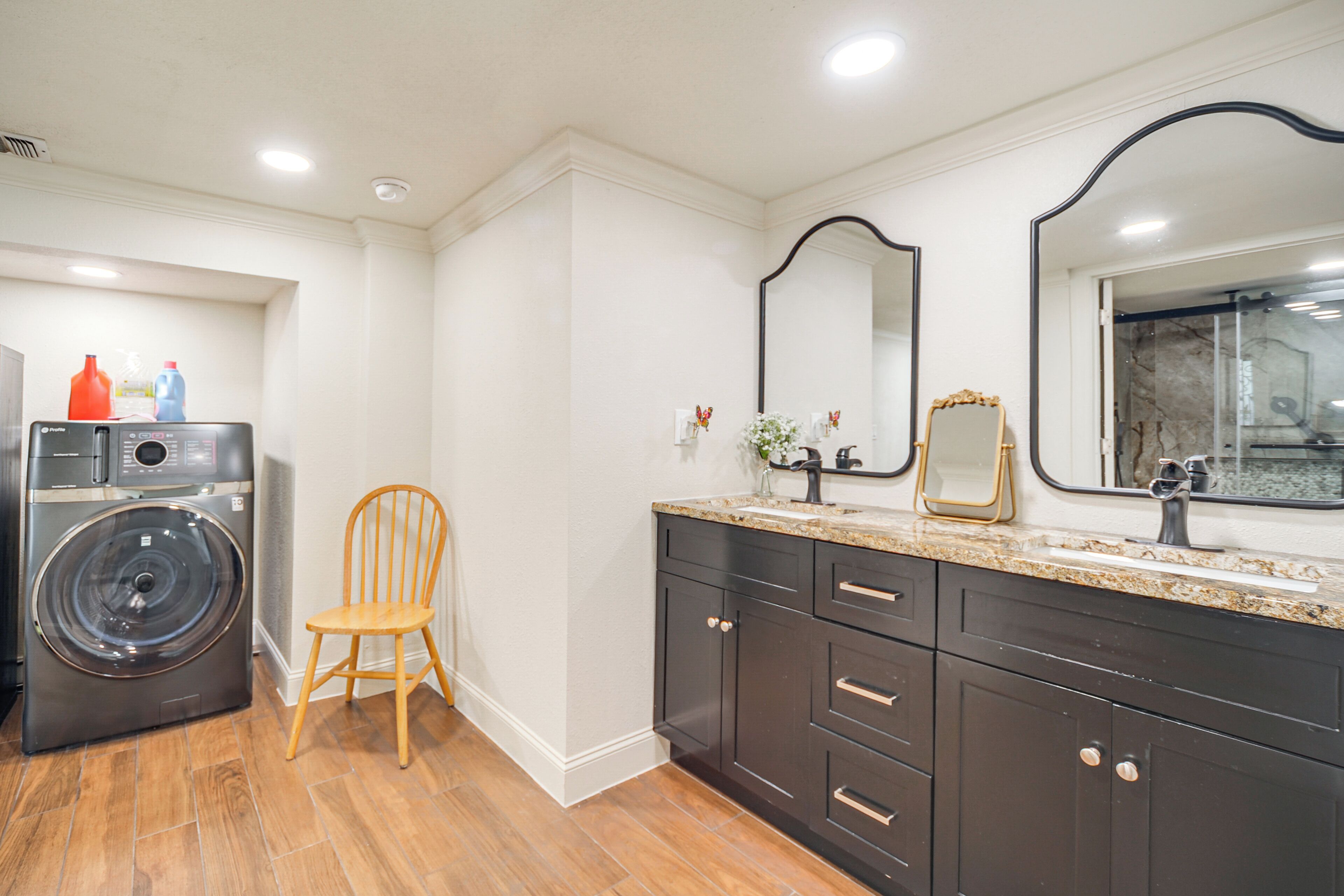 A modern bathroom with a washing machine, a wooden chair, dark cabinets, and a large mirror.