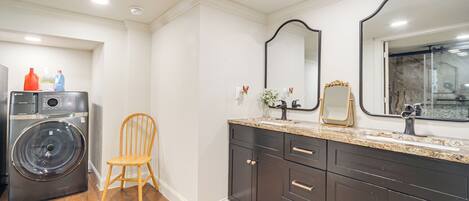 A modern bathroom with a washing machine, a wooden chair, dark cabinets, and a large mirror.
