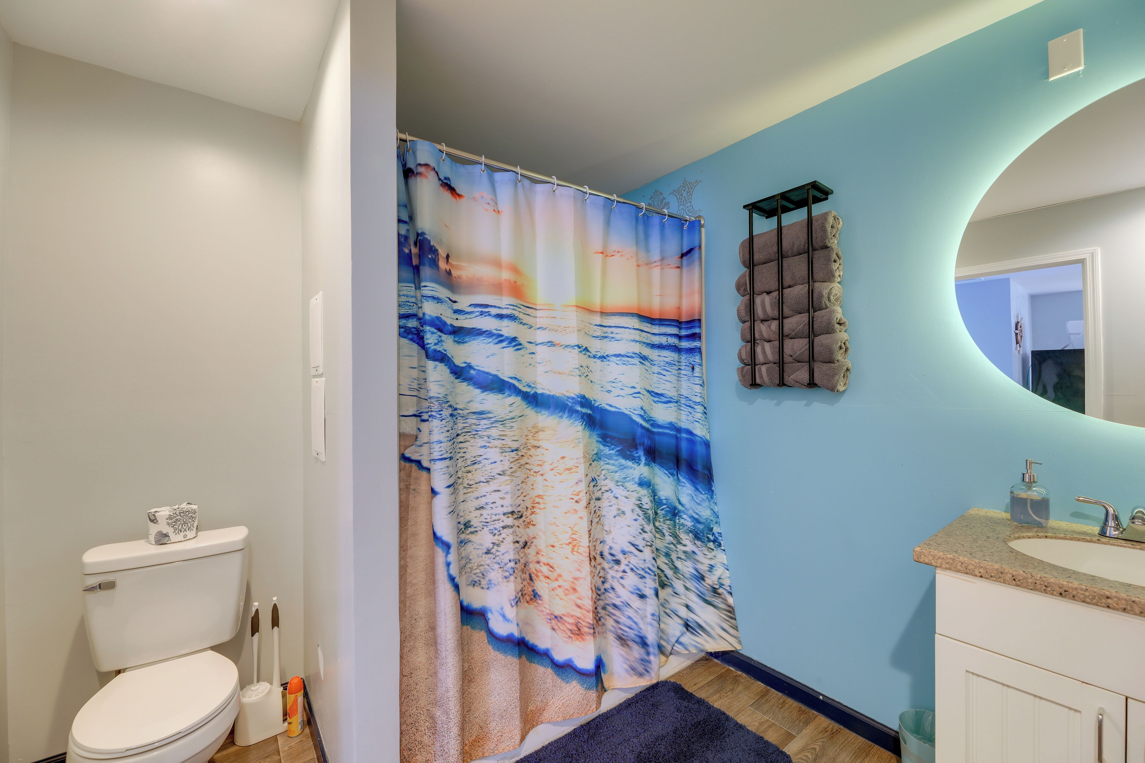 A bathroom with a white toilet, a shower curtain with a beach scene, a towel rack, and a sink with a soap dispenser.
