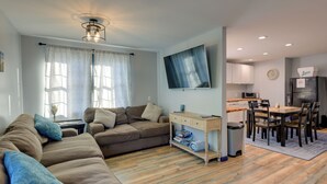 Ein Wohnzimmer mit einer Couch, einem Couchtisch und einem an der Wand montierten Fernseher. Es gibt eine Küchenzeile mit Schränken, einem Kühlschrank und einer Mikrowelle.