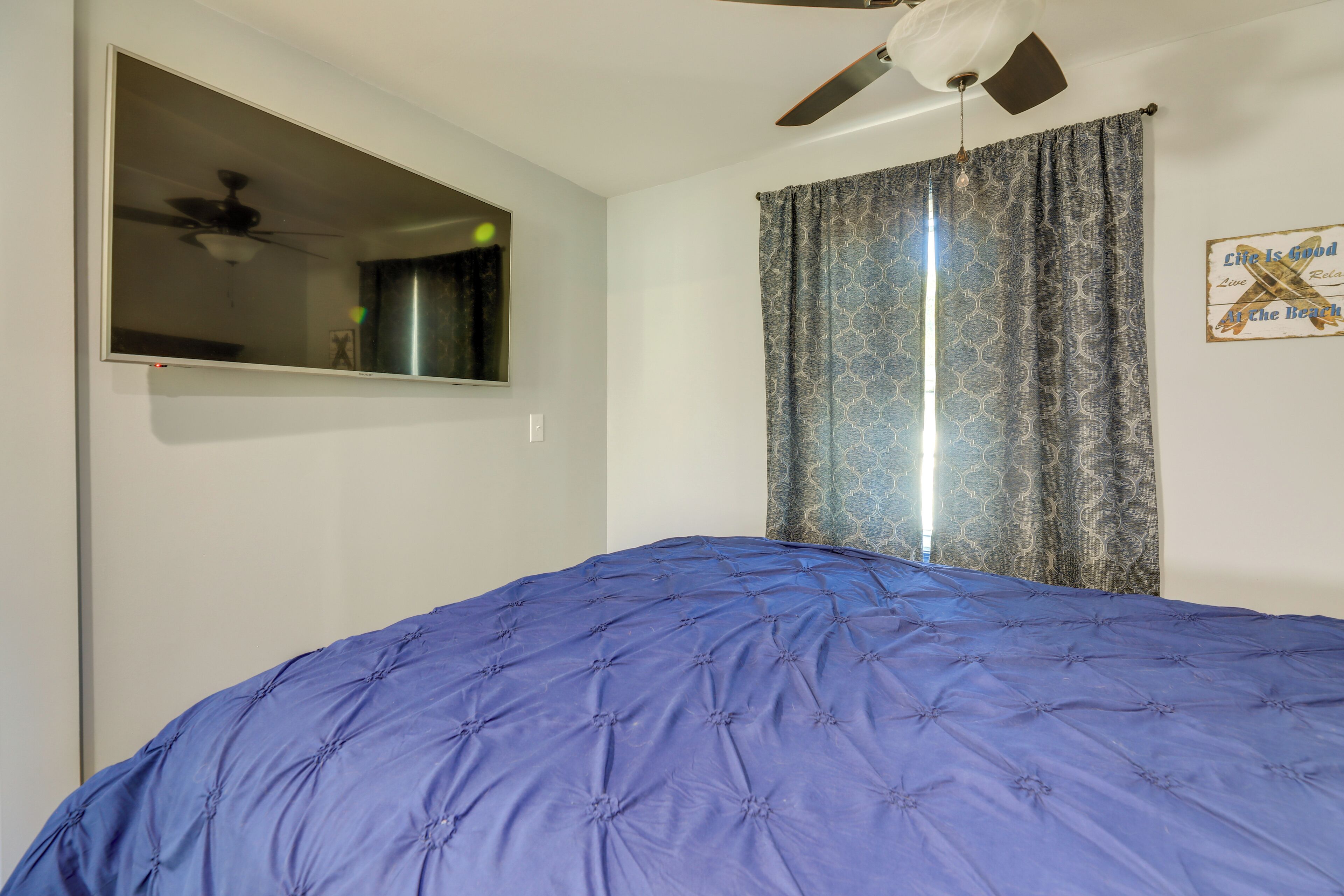 A bedroom with a large bed, a ceiling fan, a window with patterned curtains, and a wall-mounted mirror.