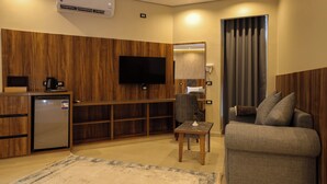 Een kleine hotelkamer met kitchenette, flatscreen-televisie, een bank, een tafeltje en een groot raam met gordijnen.