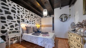 Une chambre avec un mur en pierre, un lit recouvert d’une couvre-lit à motifs floraux, une table de chevet avec une lampe et une commode avec des tiroirs.