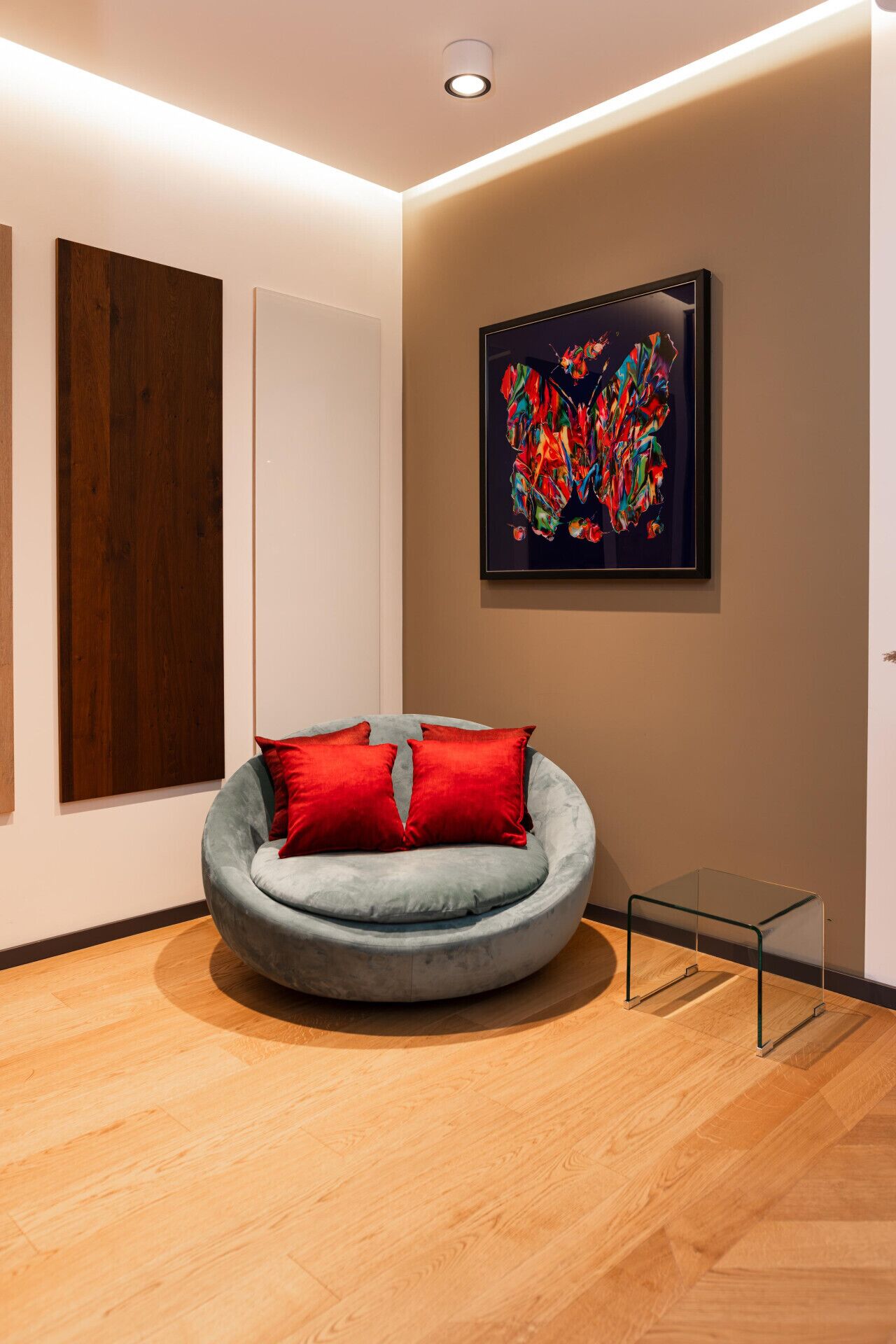 Een moderne kamer met een ronde, grijs bekleden fauteuil met rode kussens, een houten vloer, een groot abstract schilderij aan de muur en een kleine glazen tafel.