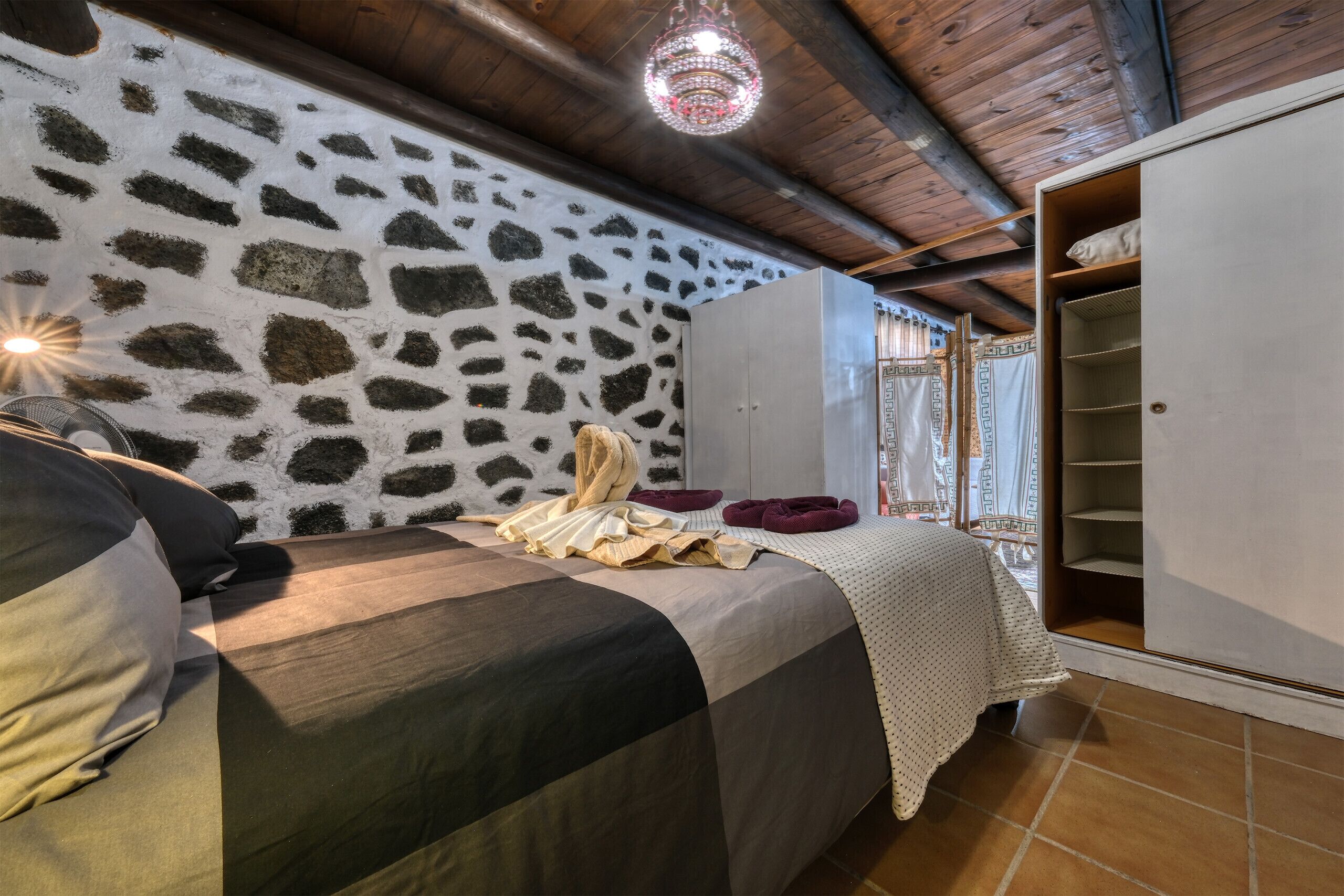 Een slaapkamer met een stenen muur, een bed met een gestreept dekbed, een kledingkast en een kroonluchter.
