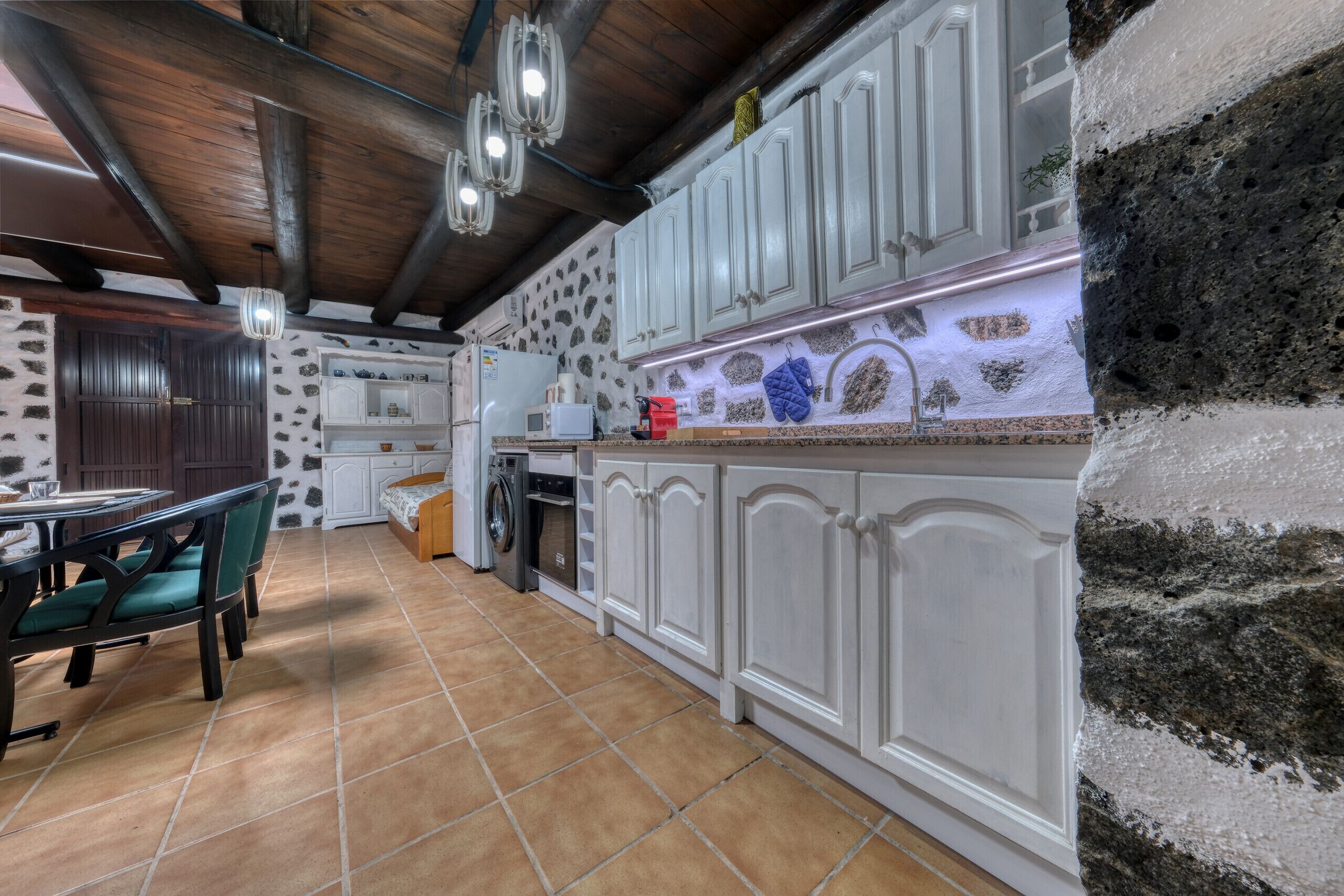 Una cocina con armarios blancos, pared de piedra y suelo de baldosas.