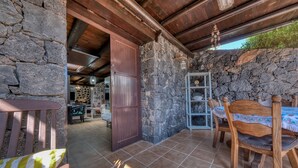 Paredão rústico de pedra, mesa e cadeiras de madeira numa área externa coberta.
