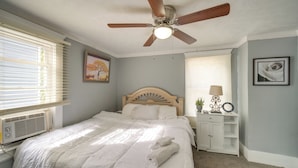 Un dormitorio con cama, ventilador de techo, ventana con persianas, mesita de noche con lámpara y un cuadro en la pared.