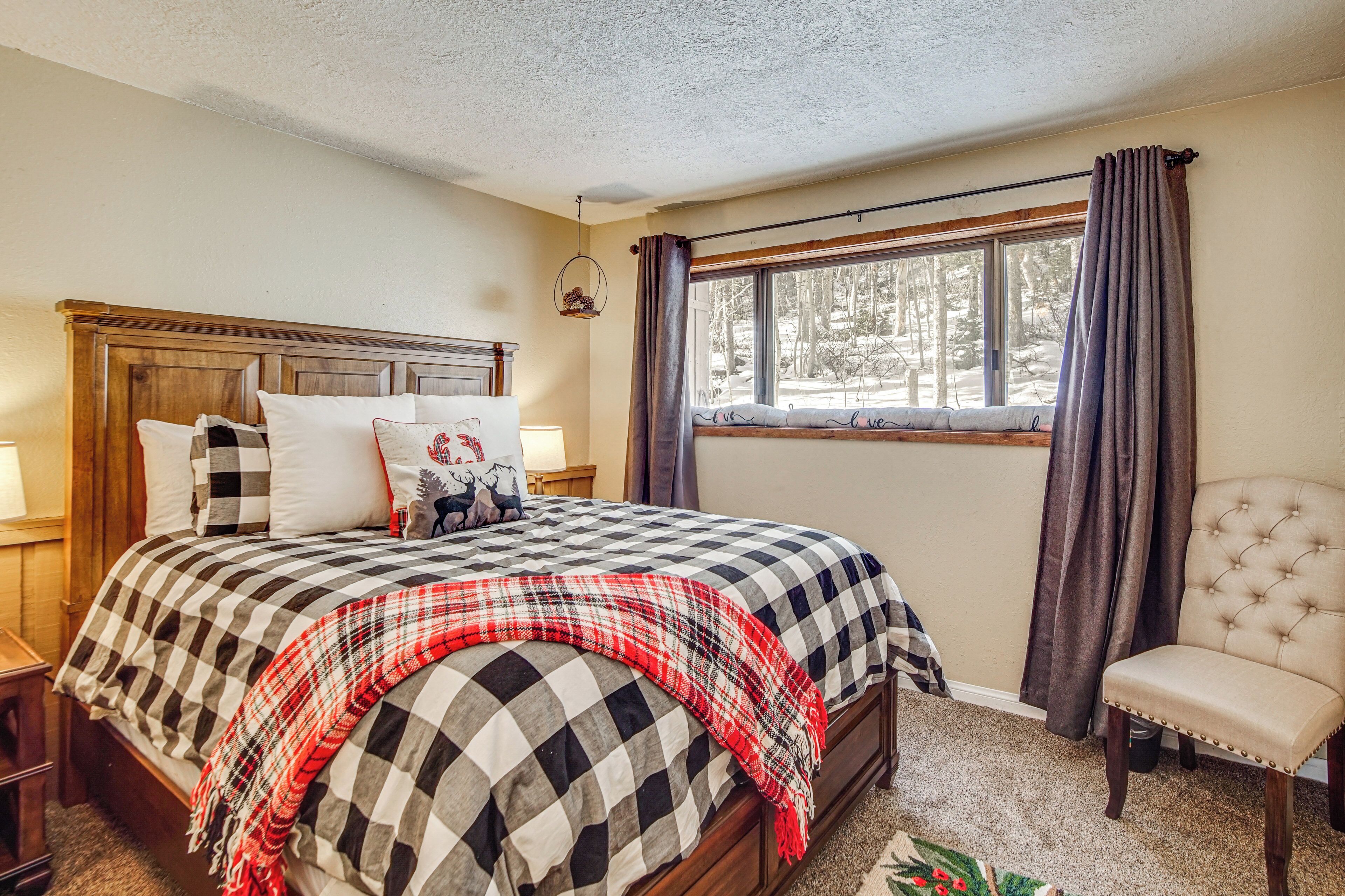 A bedroom with a wooden headboard, a checkered bedspread, a red and white plaid throw blanket, a window with curtains, and a tufted armchair.