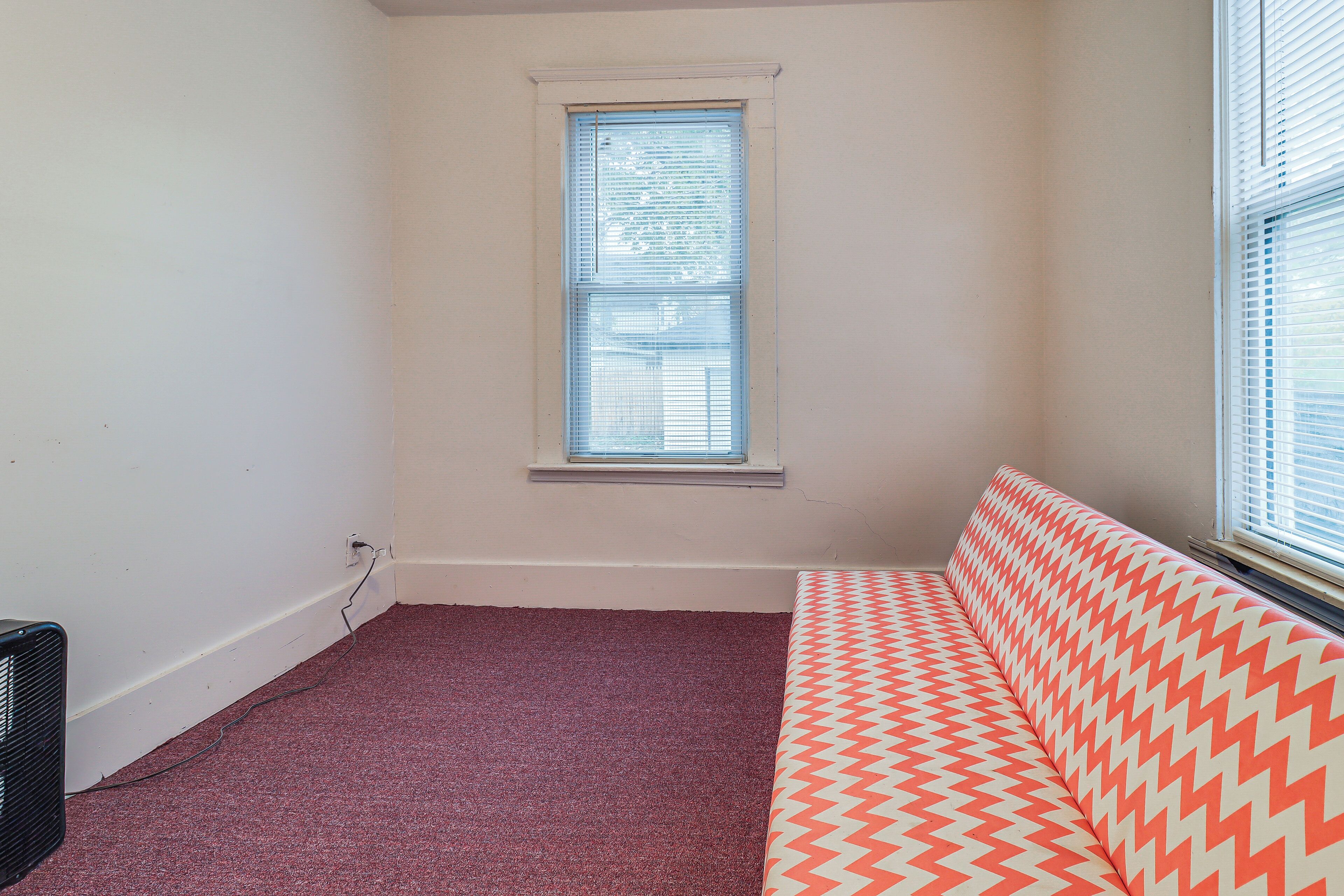 Ein Zimmer mit Fenster, Heizkörper, rotem Teppich und einem gemusterten Sofa.
