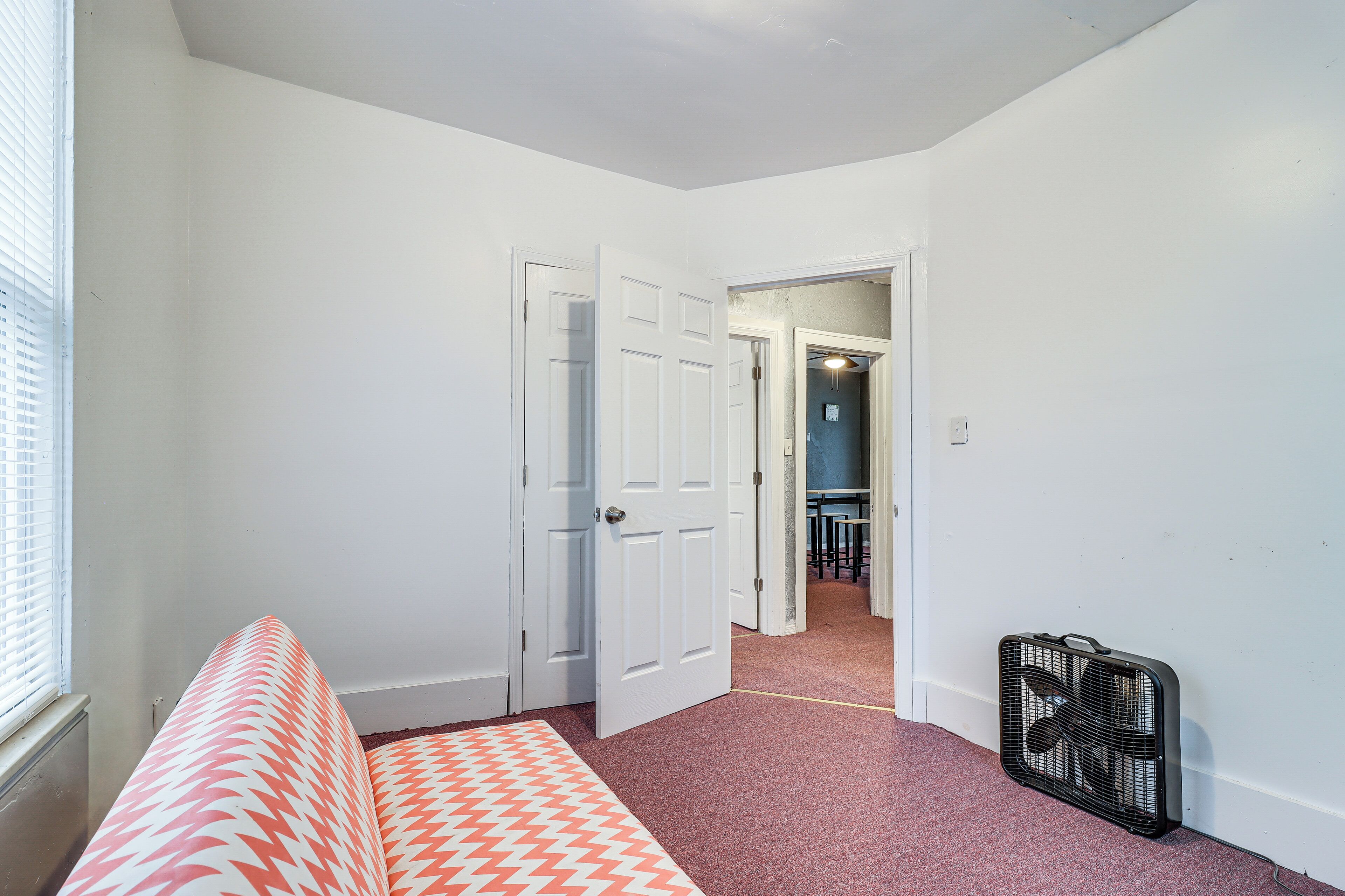A bedroom with a bed, white walls, a door, and a hallway leading to a dining area.